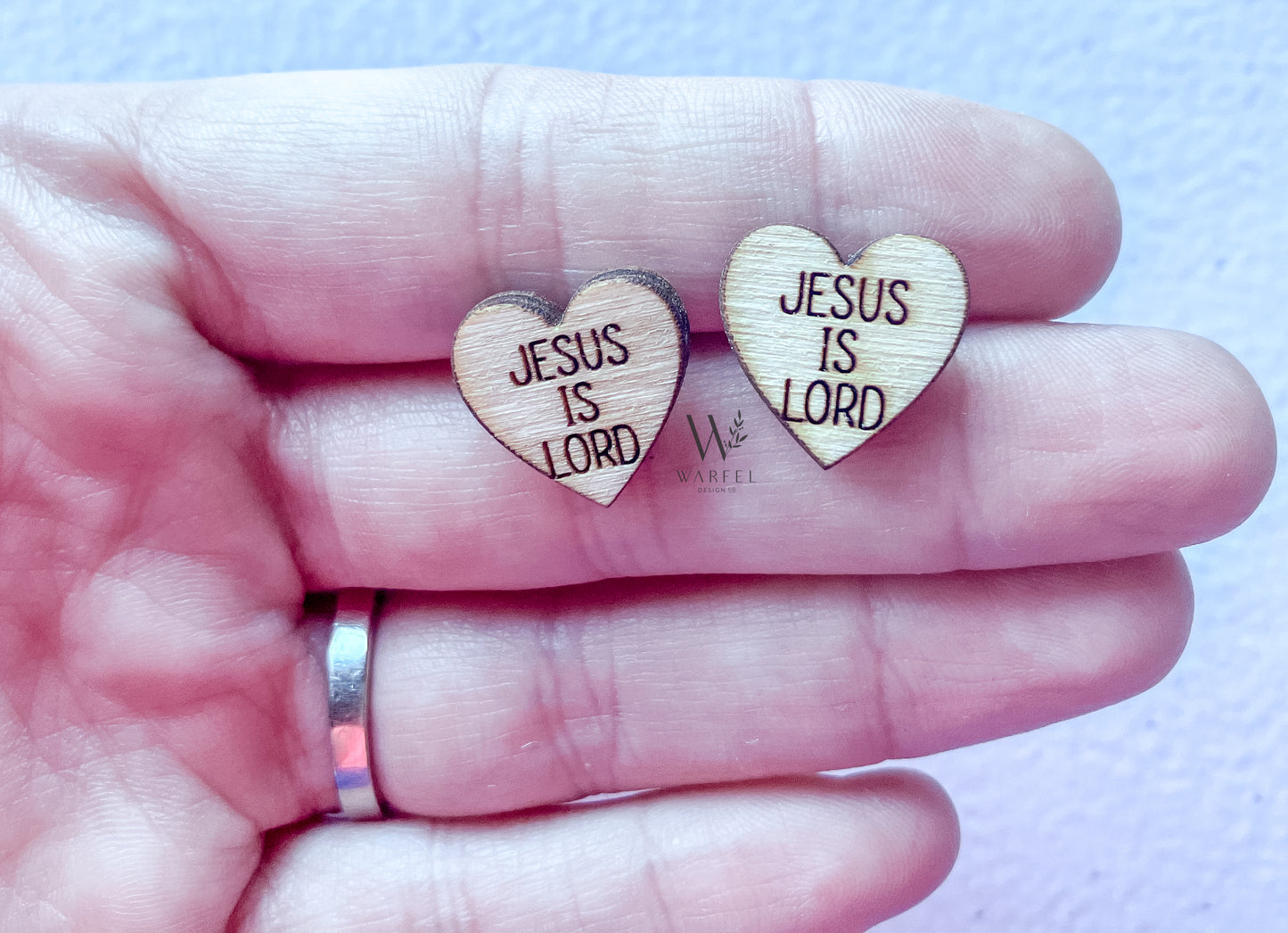 A pair of wooden stud earrings with the inscription 'JESUS IS LORD' on them, held between fingers.