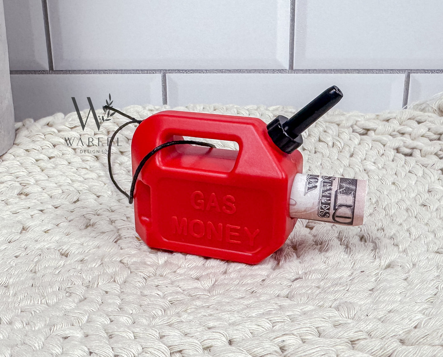 A red gas can ornament with a black spout and handle, sitting on a beige textured surface, labeled 'GAS MONEY' on its side.