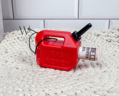 A red gas can ornament with a black spout and handle, sitting on a beige textured surface, labeled 'GAS MONEY' on its side.