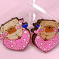 A pair of wooden pug heart stud earrings, hand-painted and coated with resin, featuring pink color with blue and white accents, and butterfly backs.
